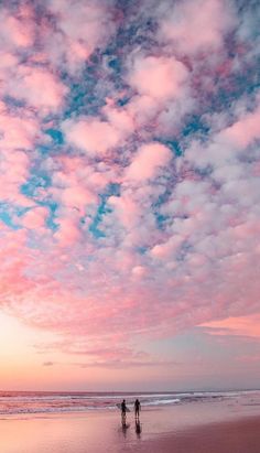 two people walking on the beach at sunset with clouds in the sky and one person holding a surfboard