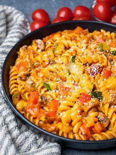 a skillet filled with pasta and vegetables on top of a table next to tomatoes