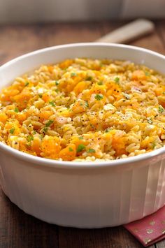 a white bowl filled with rice and vegetables on top of a wooden table next to a spoon