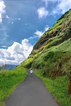 a person walking down a road near a lush green hillside on a partly cloudy day