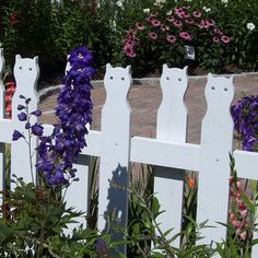 a white picket fence with purple flowers in the foreground and an orange cat statue behind it