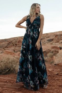 a woman standing in the desert wearing a black and blue floral print dress with her hands on her hips