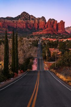 an empty road in the desert with mountains in the backgrouds and trees on either side