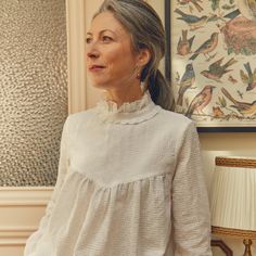 an older woman standing in front of a wall with birds on it and a lamp next to her