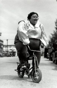 an old photo of a woman riding a bike down the street with her mouth open