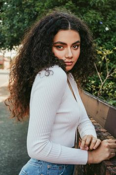 a woman with curly hair is leaning against a rail and looking at the camera while wearing a white top