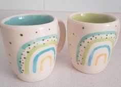 two ceramic mugs sitting on top of a counter next to each other, one with a rainbow painted on it