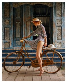 a woman in a dress and hat standing next to an old fashioned bicycle on a wooden deck