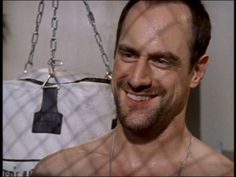 a man with no shirt smiling in front of a chain hanging from the ceiling behind him