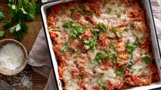 a casserole dish with meat, cheese and parsley on the side next to a bowl of grated parmesan cheese