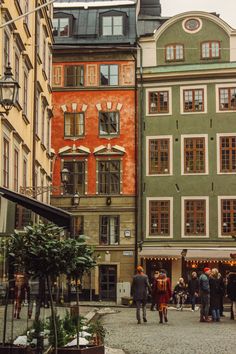 people are walking around in an old european city square with many buildings and plants on the cobblestone streets