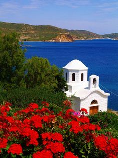 a white church with red flowers in the foreground and blue water in the background