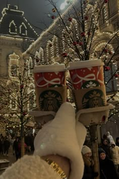 two starbucks cups are held up in front of a christmas tree with lights on it