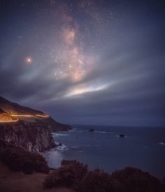 the night sky is filled with stars and clouds above an ocean side road that stretches into the distance