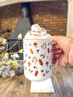 a hand holding a coffee mug with autumn leaves on it and the lid covered in whipped cream