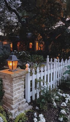 a white picket fence with a light on it in front of some flowers and trees