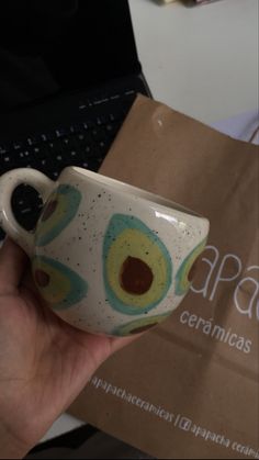 a hand holding a coffee cup with an avocado design on it and a laptop in the background