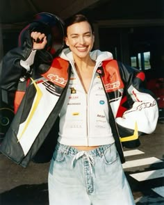 a woman is standing in front of a car and holding her jacket over her shoulders