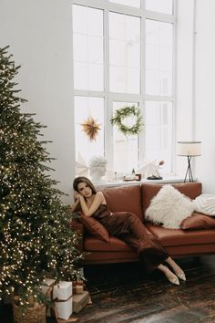 a woman sitting on a couch next to a christmas tree in a room with large windows