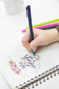 a person holding a pen and writing on a notepad with colored pencils next to it