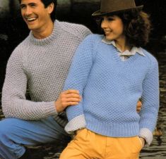 two people in sweaters and hats are posing for a photo by the water with their arms around each other