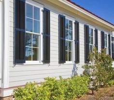 a white house with black shutters on the front and side windows, along with shrubbery