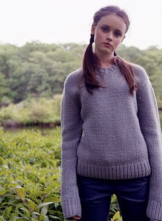 a woman standing in front of bushes wearing a gray sweater and blue jeans with her hands in her pockets