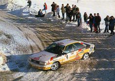 a rally car driving down a snow covered road with people standing on the side of it