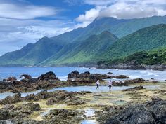 two people are walking on the rocks near the water with mountains in the back ground