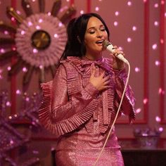 a woman singing into a microphone in front of a pink wall with lights on it