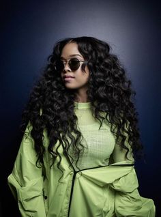 a woman with long curly hair wearing sunglasses and a green top is standing in front of a dark background