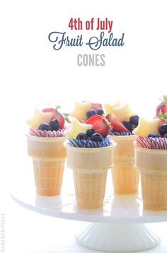 four ice cream cones filled with fruit on top of a white cake platter that says 4th of july fruit salad cones