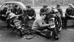 a group of men sitting on the side of a road next to parked motor cycles