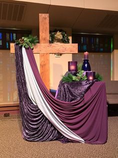 the cross is decorated with purple and white cloths, surrounded by greenery as well as candles