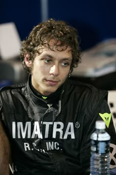 a man with curly hair sitting next to a water bottle and looking at the camera