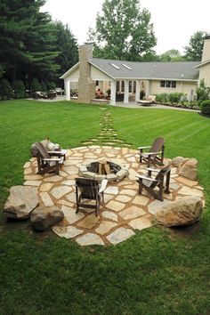 an outdoor fire pit with chairs around it in the middle of a grassy area next to a house