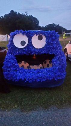 an inflatable blue monster sitting on top of a lush green field next to parked cars