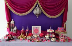 a table topped with cakes and desserts under a purple drape covered wall next to a window