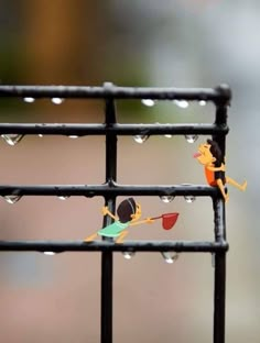 two small figurines are on top of a metal fence with water droplets hanging from them