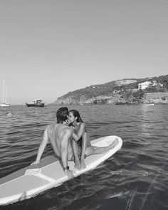 two people on a surfboard kissing in the water with boats in the back ground