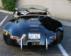 an old black sports car parked in a parking lot next to a wall and bushes