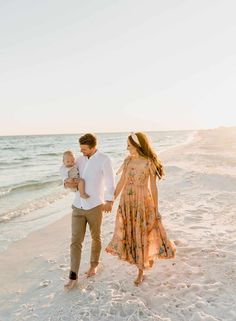 a man and woman walking along the beach with a baby in their arms as the sun sets