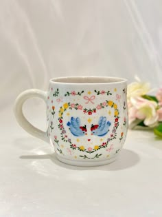 a cup with two birds painted on it next to some flowers and a white background