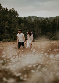 a man and woman holding hands walking through tall grass in front of some pine trees