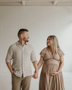 a pregnant couple holding hands and smiling at each other in front of a white wall