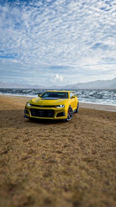 a yellow sports car is parked on the beach near the water and clouds in the sky