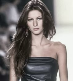 a woman with long brown hair walking down a runway wearing a black leather dress and earrings