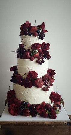 a three tiered white cake with red flowers and berries on the top is sitting on a wooden table