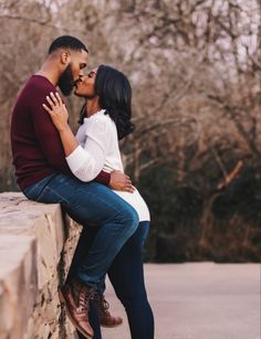 a man and woman sitting on a stone wall with trees in the background, kissing