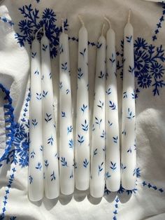 six candles are lined up on a tablecloth with blue and white flowers in the background